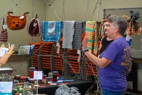 Two women stand near a display of knitted items and jars at an indoor exhibition.