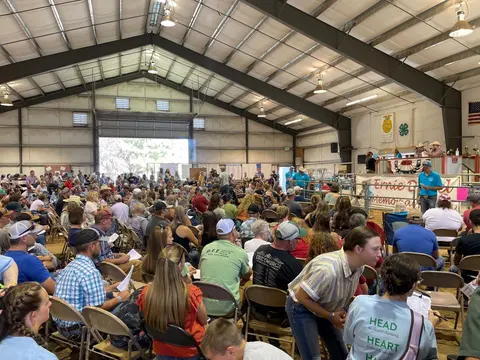 A large crowd gathers inside a metal-framed agricultural arena or livestock pavilion.
