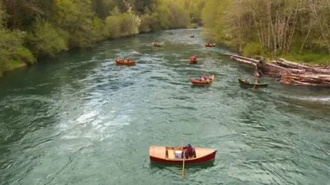 Several boats navigate down a river surrounded by forested banks.