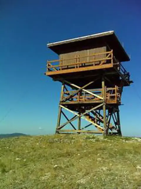 A wooden fire lookout tower stands on a grassy hilltop against a clear blue sky.