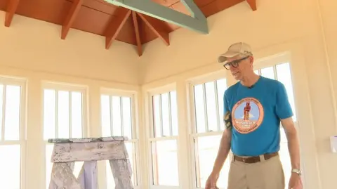 A man stands inside a sunlit room with multiple windows and a wood-beamed ceiling.