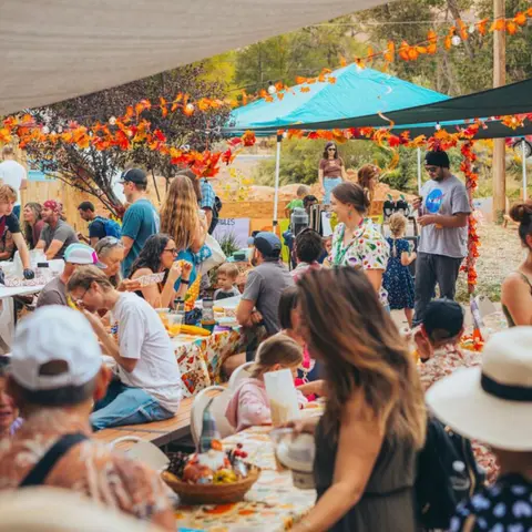 A crowd of people gathers outdoors around tables decorated for autumn.