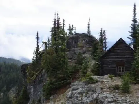 A small log cabin sits on a rocky ridge among evergreen trees.