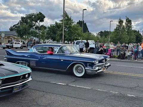 A blue vintage car driving down a street with people watching on the sidewalk.