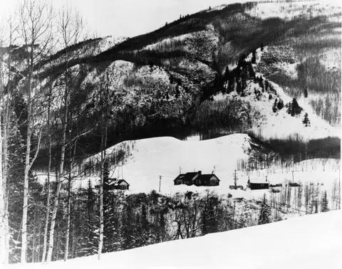 A black and white photograph showing a few small buildings nestled in a snowy mountain landscape.