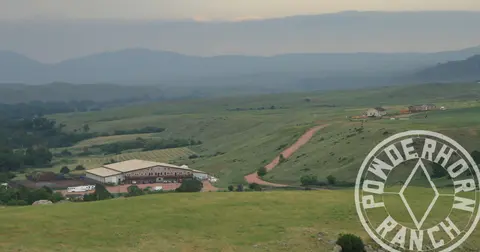 A wide landscape view of a ranch with green hills and mountains in the background.