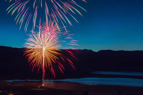Colorful fireworks explode in the night sky above a landscape with water and hills.