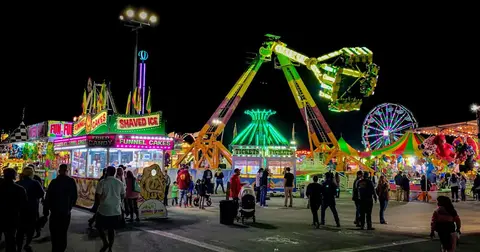 A brightly lit carnival at night with rides, food booths, and visitors.