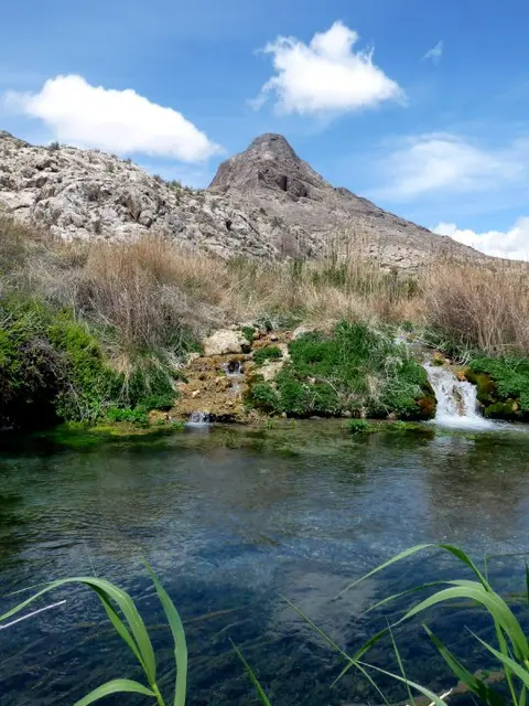 A natural spring with small waterfalls flows into a clear pool beneath a rocky mountain.