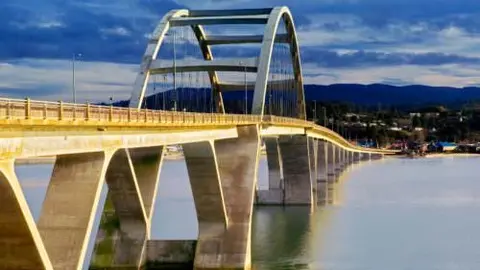 A large arch bridge spans across a body of water under a cloudy sky.