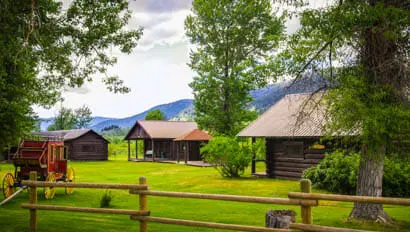 A view of rustic log cabins and a vintage wagon in a grassy field with mountains in the background.