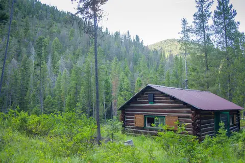 A log cabin sits in a lush green valley surrounded by a dense forest and mountains.