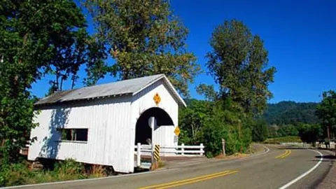 A white covered bridge sits beside a paved road surrounded by trees and mountains under a blue sky.