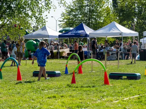 A crowd of people enjoys an outdoor event with tents and agility equipment on a grassy field.