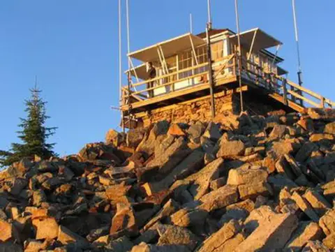 A fire lookout tower perched on a rocky summit against a clear blue sky.