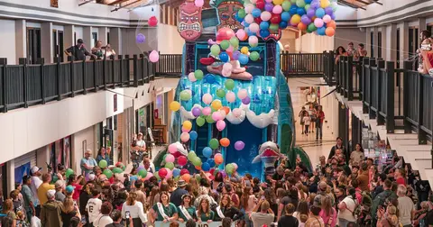 A crowd of people gathers indoors around a large inflatable slide decorated with balloons.