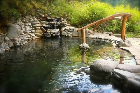 A natural hot spring pool with stone walls, a small waterfall, and a wooden handrail.