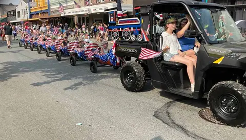 People riding in a decorated utility vehicle and wagons during a parade on a street.