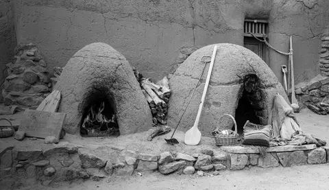 Two adobe ovens stand outside a textured wall with baking tools and firewood.