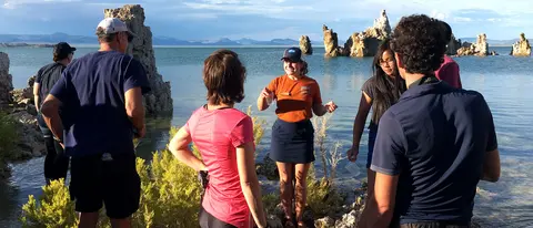 A guide speaks to a group of visitors standing near a lake with rock formations.