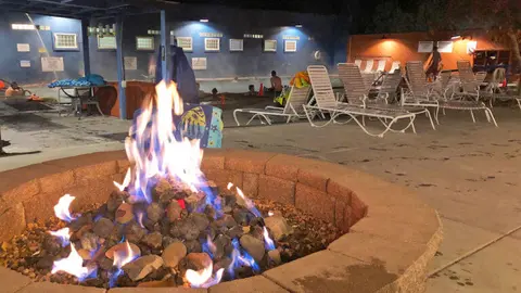 A stone fire pit burns brightly on a patio next to a pool at night.