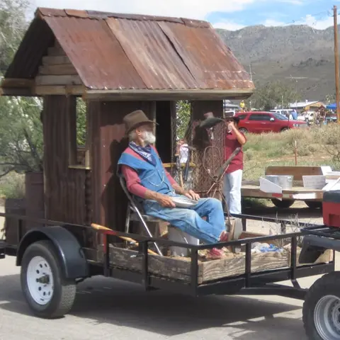A man in a cowboy hat sits on a trailer holding a small structure.
