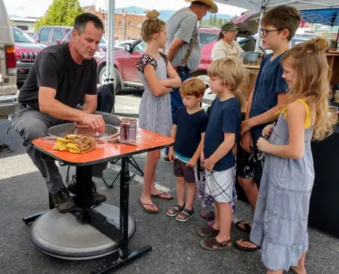 A man demonstrates pottery making on a wheel for children outdoors.