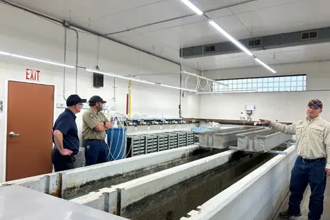 Three men stand inside an industrial or hatchery building with large concrete water troughs.