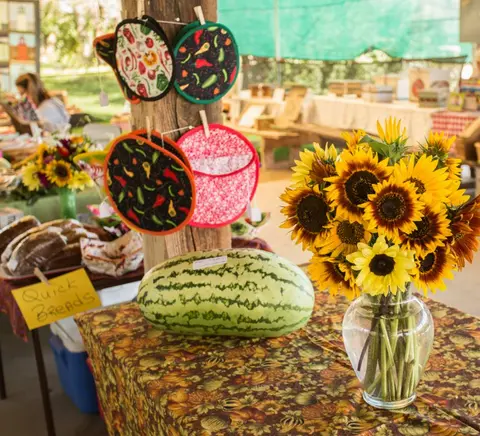 A table at a market features a large watermelon, a vase of sunflowers, and hanging pot holders.