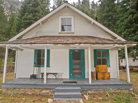 A historic white wooden house with a porch and teal trim sits among trees.