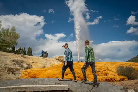 Two people walk near an erupting geyser against a scenic sky.