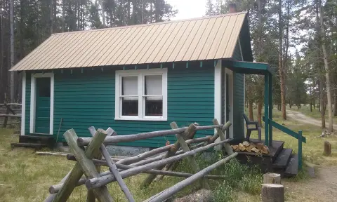 A small green cabin stands in a wooded area with a rustic wooden fence in the foreground.