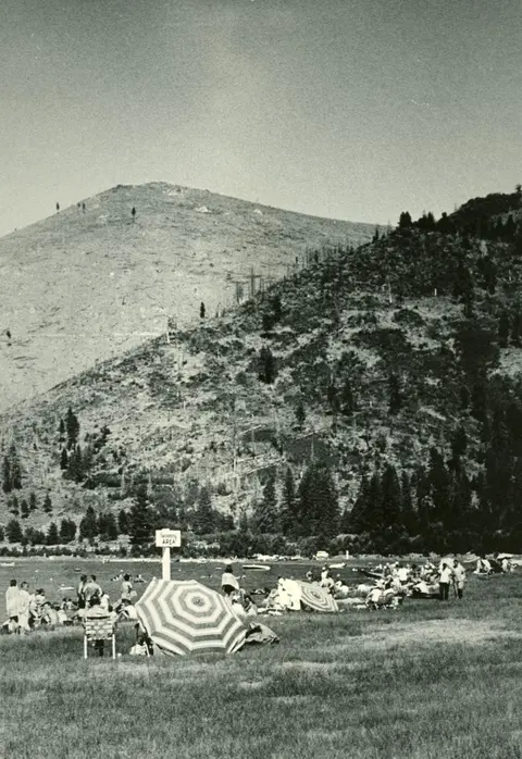 Black and white historic photo showing people relaxing on a grassy shore by a lake with a large mountain in the background.