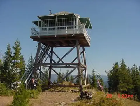 A fire lookout tower stands atop a wooded mountain ridge under a clear sky.