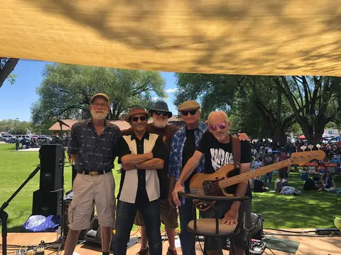 A band of five older men pose on an outdoor stage with an audience in the background.