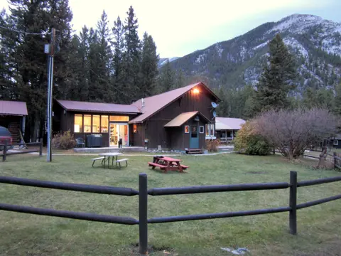 A rustic wooden cabin sits in a clearing surrounded by mountains and pine trees.