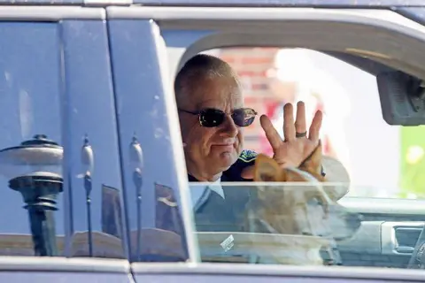A person wearing sunglasses waves from inside a vehicle alongside a dog.