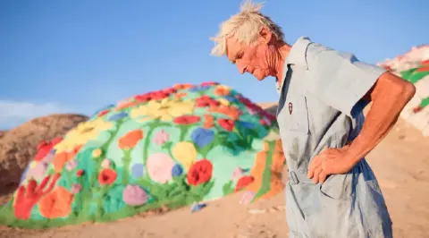 An older man stands near a brightly painted outdoor art structure.