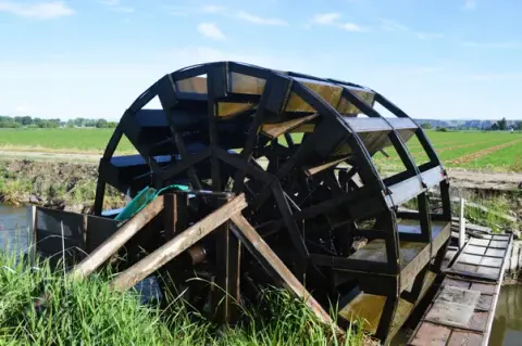 A large wooden water wheel sits beside a small stream in a grassy field.