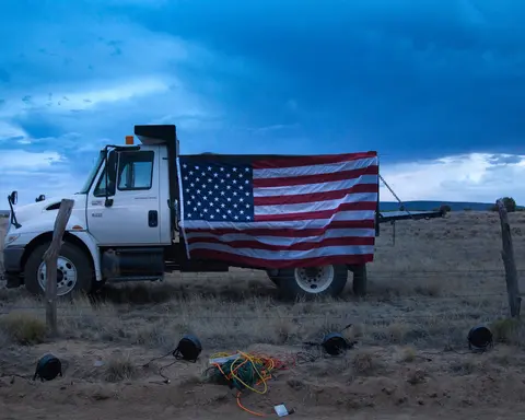A large American flag hangs on the side of a white truck parked in a field.