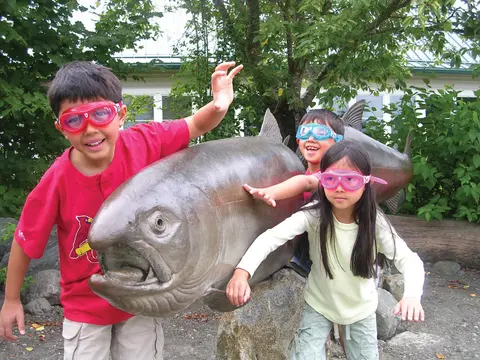 Three children wearing swim goggles pose with large fish sculptures.