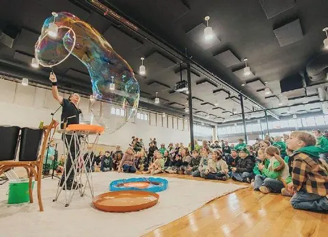 A performer blows a giant bubble for a large group of children indoors.