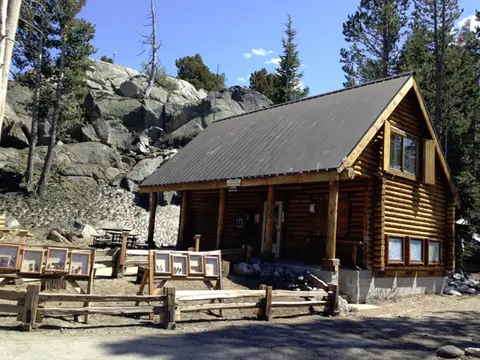 A rustic log cabin sits in a mountainous, rocky outdoor setting.