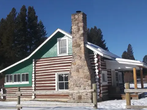 A log cabin with a stone chimney stands in a snowy landscape under a clear blue sky.
