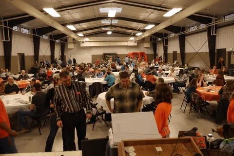 A crowd of people gathering inside a large hall with tables and chairs.