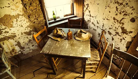 A weathered wooden table with hats and a bottle sits in a rustic room with crumbling walls.