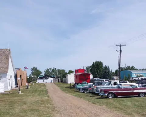 An outdoor scene featuring classic cars, a red caboose, and historic buildings along a dirt path.