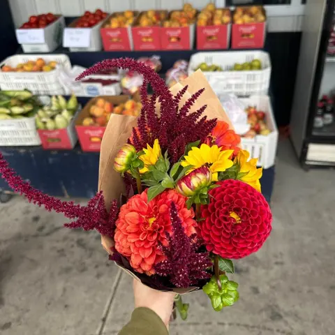 A person holds a colorful bouquet of flowers in front of a market display of produce.