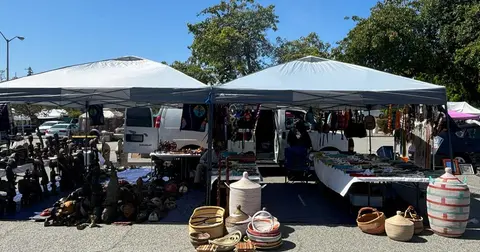 An outdoor market booth displays various crafts, statues, and woven baskets under tents.