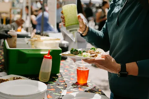A person squeezes green sauce onto tacos at a food stall.